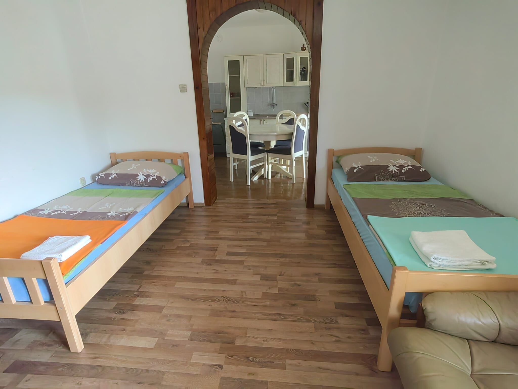 A room with two single beds, a wooden floor, a dining area with a table and chairs, and a kitchen in the background.