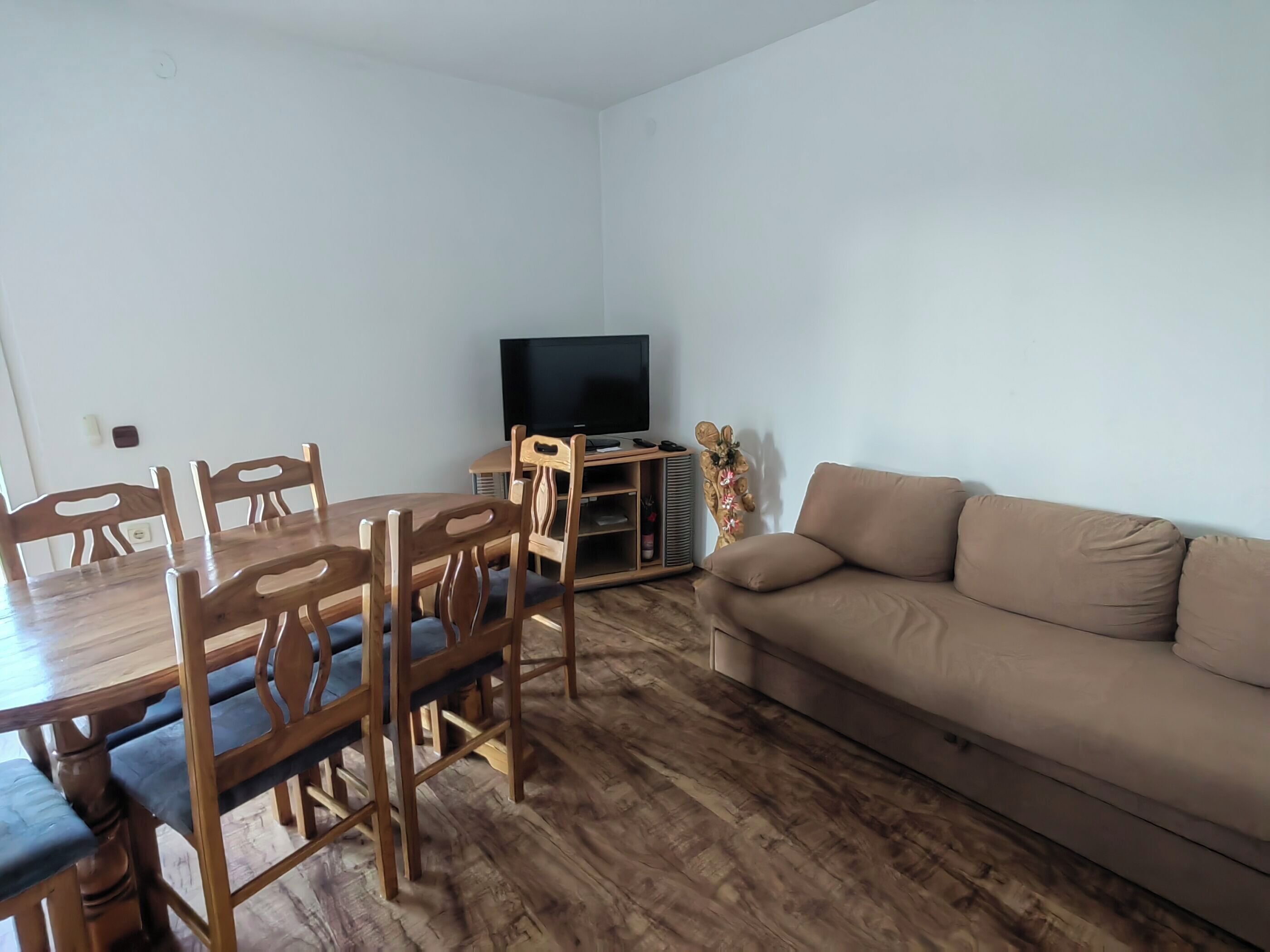 A living room with a wooden dining table, chairs, a beige sofa, a flat-screen TV, and a decorative plant.