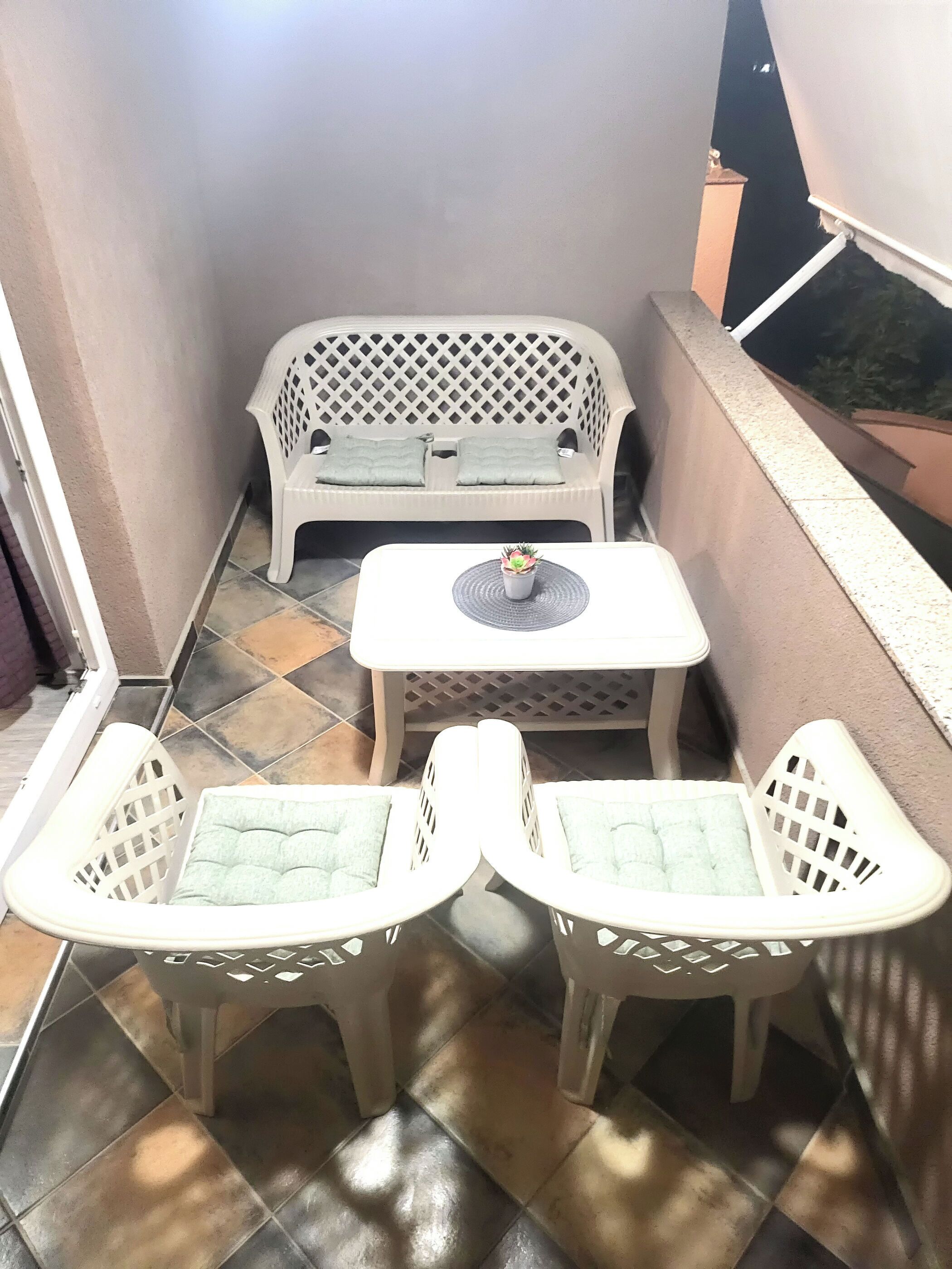 A small outdoor seating area with a white plastic sofa and two white plastic chairs, a small table, and a potted plant.