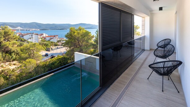 A balcony with a glass barrier overlooking a pool and a view of a coastal town. - Villa Delux View (Slivno)
