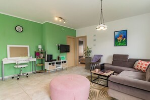 A living room with a green wall, a brown sofa, a pink ottoman, a coffee table, a TV, and a desk with a computer and shelves. - Villa May (Tinjan)