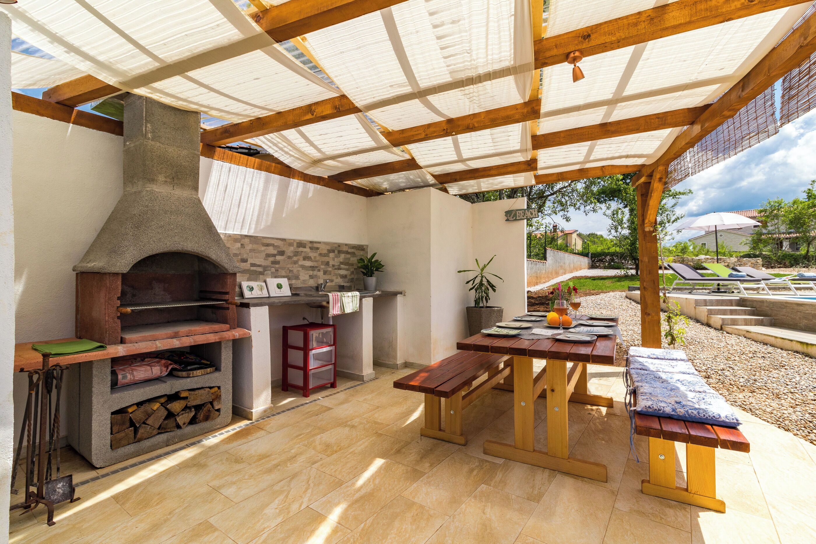 Een terras buiten met een grill van natuursteen, houten banken en een tafel met uitzicht op de tuin.