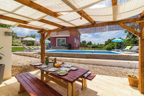 A wooden pergola with a pool and a table set for dining, overlooking a swimming pool and a house. - Villa May (Tinjan)