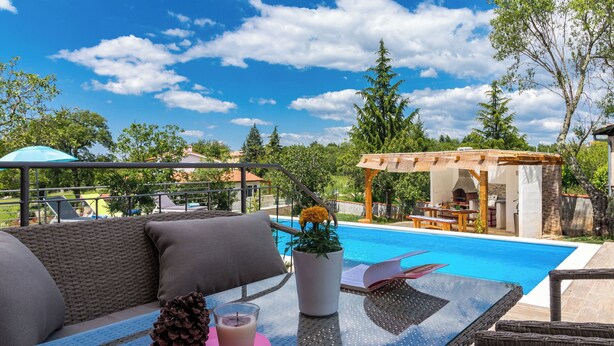 Ein Poolbereich mit Korbuschrohsofa, Glastisch und einer Pergola mit Grill.