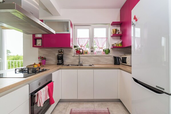 A modern kitchen with white and pink cabinets, a stainless steel range, and a window with a view of greenery. - Villa May (Tinjan)
