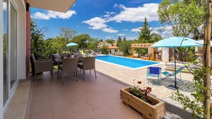 Ein Poolbereich mit Tisch und Stühlen, einem Holz-Pflanzgefäß mit Blumen und Blick auf einen Swimmingpool und umliegende Gebäude.