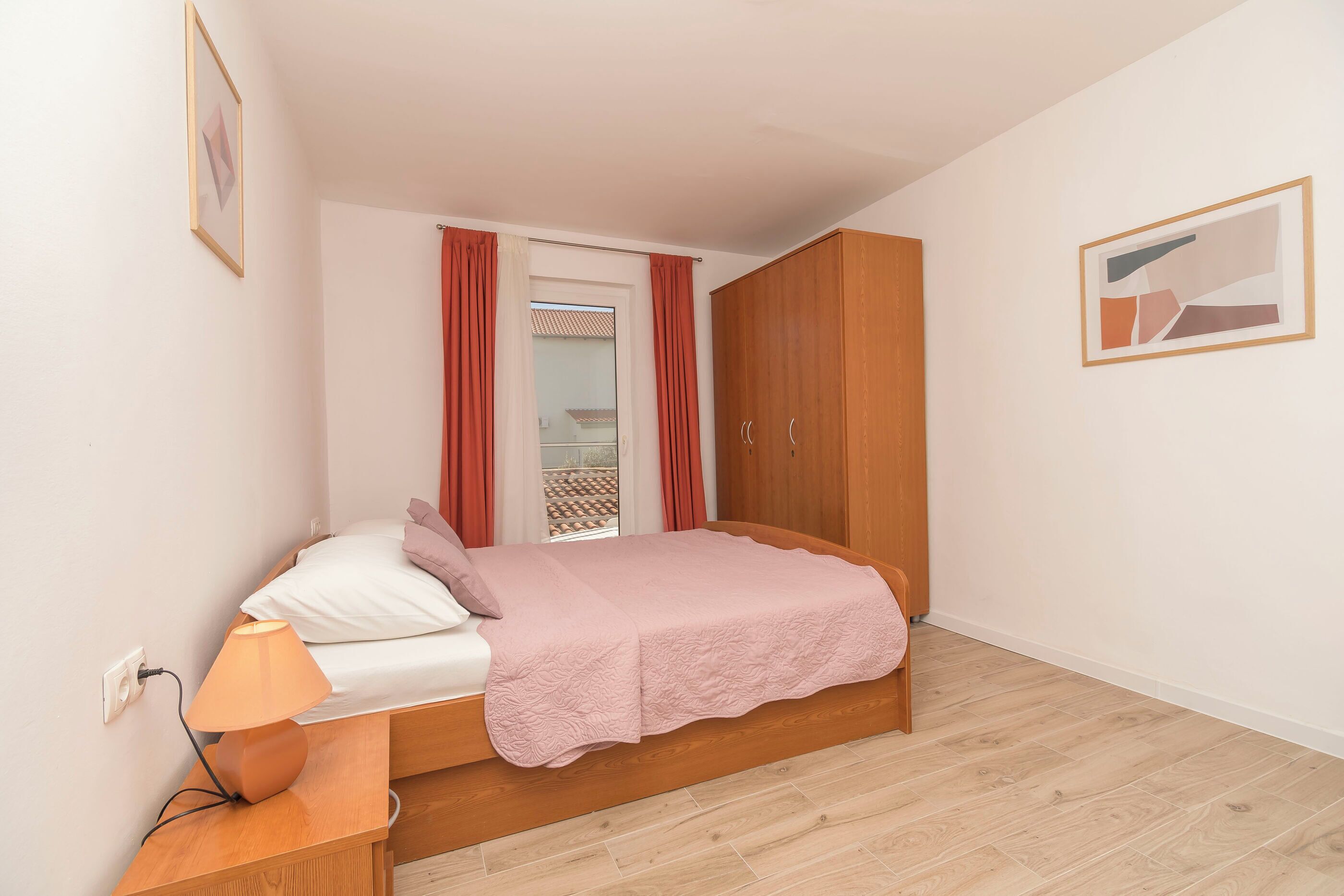 A bedroom with a bed, a nightstand, a lamp, a wardrobe, and a window with curtains.