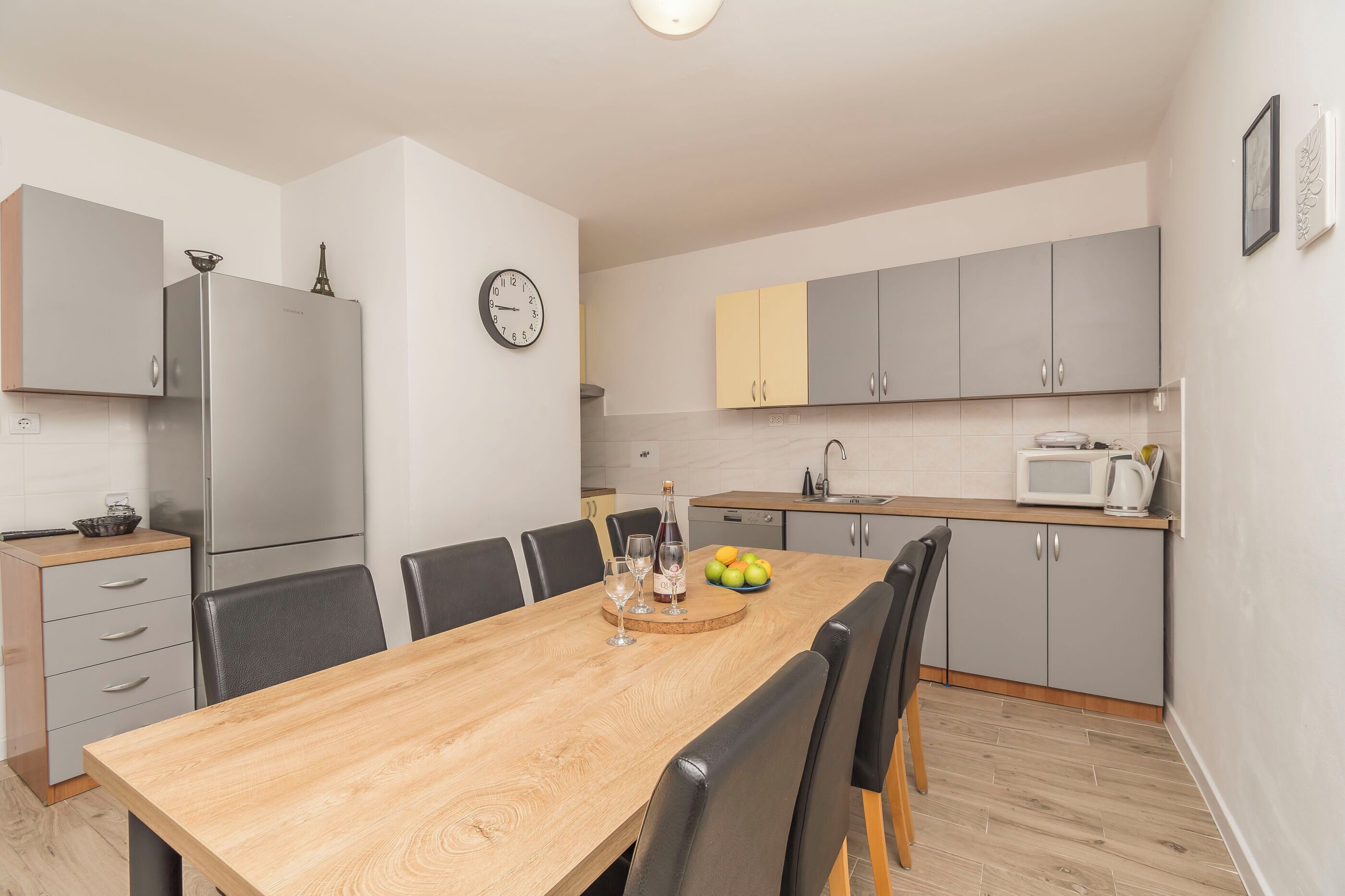 A modern kitchen with grey cabinets, a stainless steel refrigerator, a wooden dining table with chairs, and a clock on the wall.