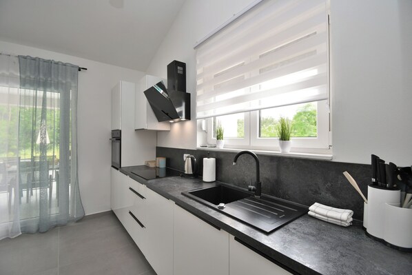 A modern kitchen with a black sink, white cabinets, and a window with blinds. - Villa Tersaz (Sveta Nedelja)
