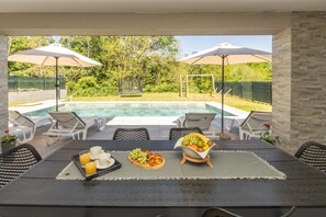 An outdoor dining area with a pool view, including a table set with fruits, a tray of orange juice, and a croissant. - Villa Tersaz (Sveta Nedelja)