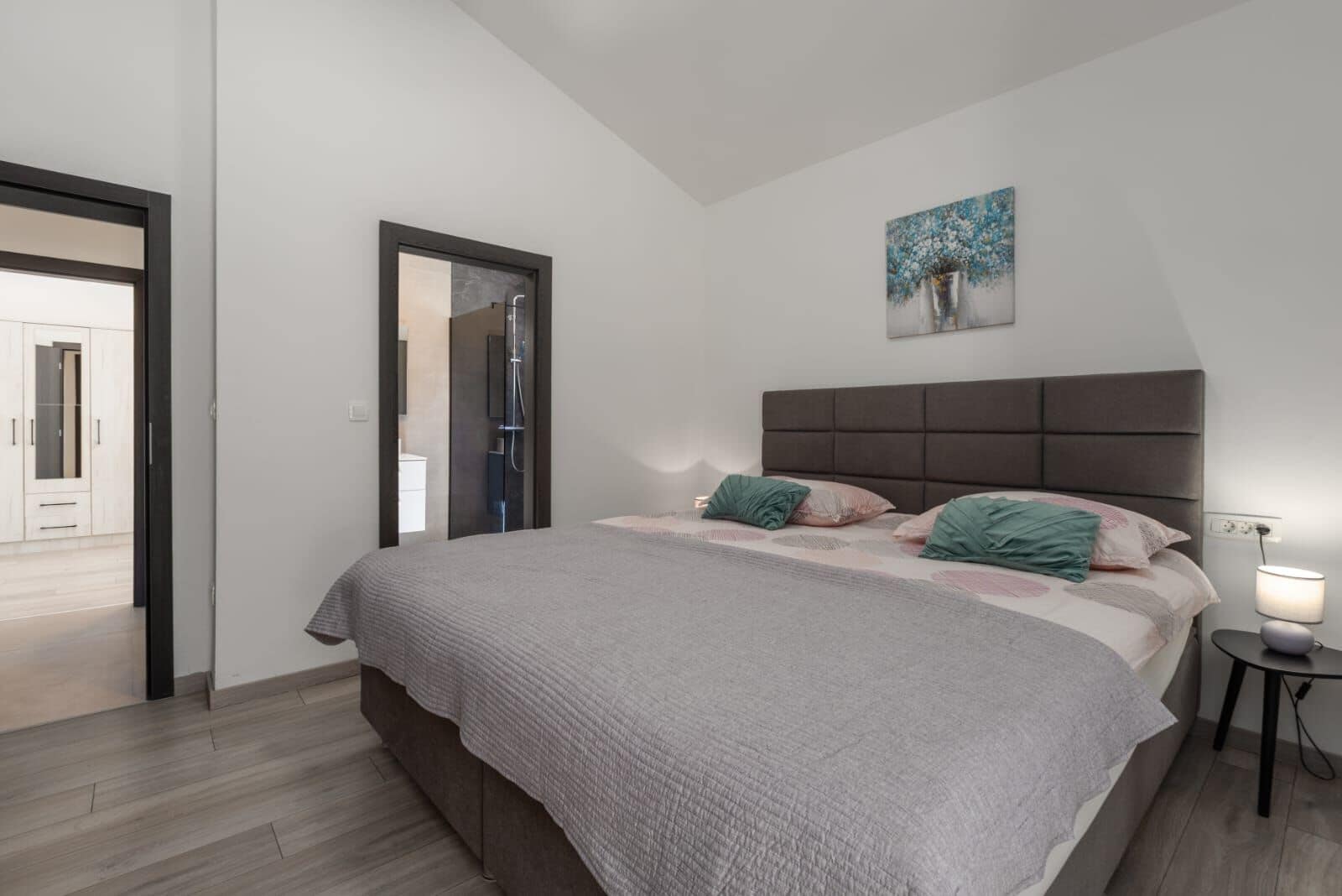 Quarto com cama, mesa de cabeceira, quadro e porta que dá para outro cômodo.