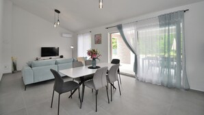 A modern living room with a light blue sofa, a dining table with four chairs, a flat-screen TV mounted on the wall, and sheer curtains on the windows. - Villa Tersaz (Sveta Nedelja)