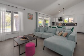 A modern living room with a sofa, a coffee table, and a pink ottoman. There is a kitchen area with a dining table and chairs visible through an open door. - Villa Tersaz (Sveta Nedelja)