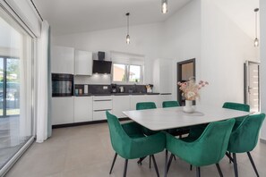 Egy modern konyha fehér szekrényekkel, fekete színű páraelszívóval, valamint egy étkezővel, melyben fehér asztal és zöld székek találhatók.