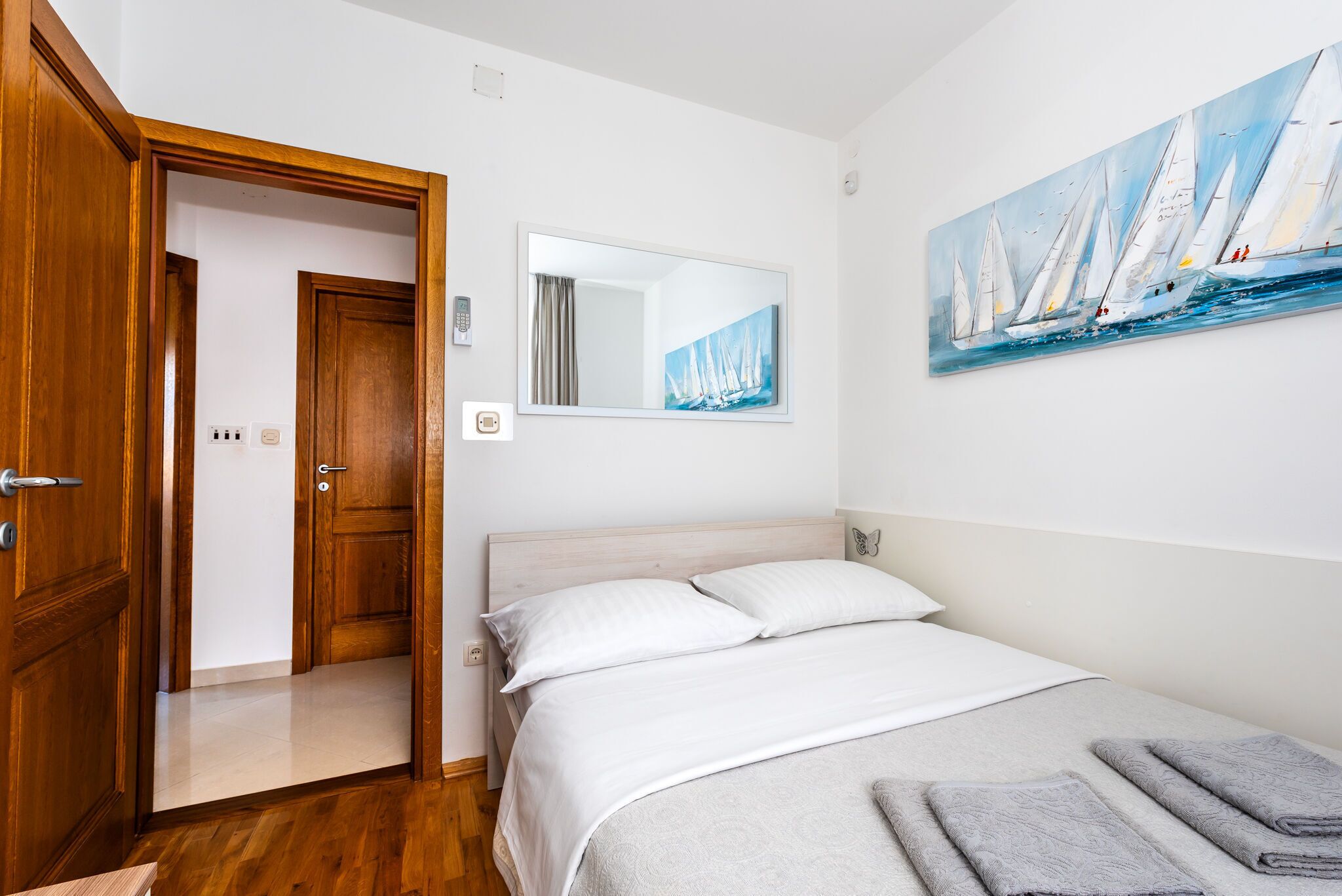 A bedroom with a wooden door, a bed with white bedding, a painting of sailboats, and a mirror on the wall.
