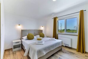 A bedroom with a bed, bedside tables, a window with curtains, and a view of greenery. - Villa St Vid 4 (Privlaka)