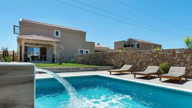 A swimming pool with a water feature, surrounded by a stone wall and a modern building with a tiled roof. - Villa St Vid 4 (Privlaka)