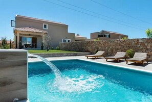 Una piscina con fuente de agua, rodeada por un muro de piedra y un edificio moderno con tejado de baldosas.