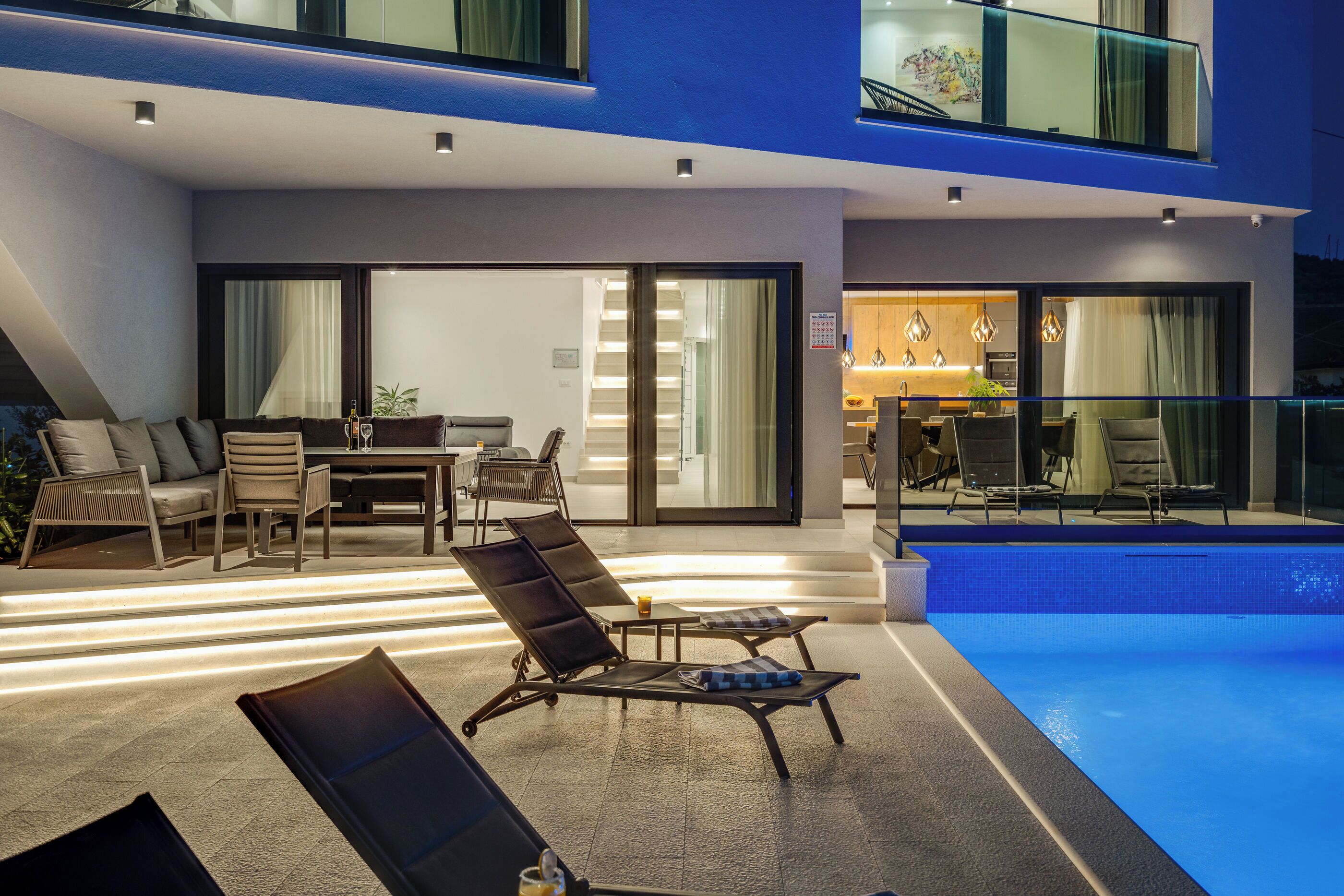 Área moderna da piscina ao ar livre, com espreguiçadeiras, uma mesa de jantar e uma cozinha visível através de portas de vidro.
