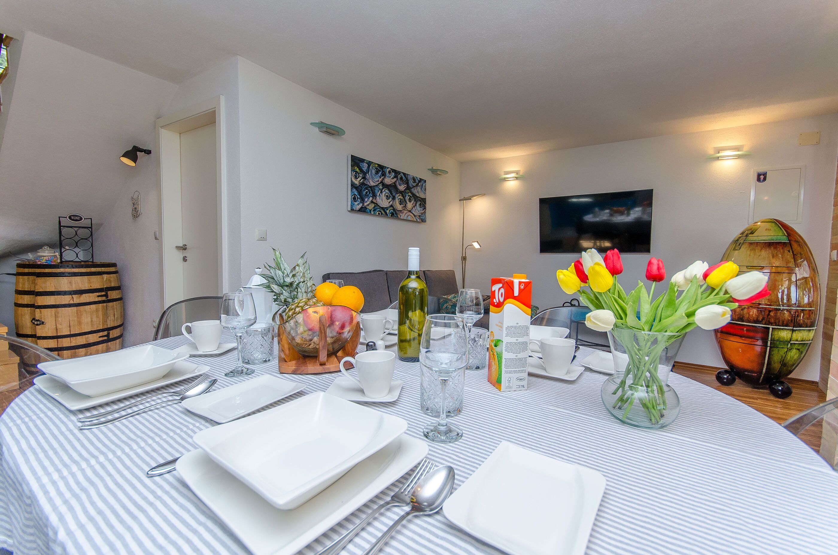 Een eetgedeelte met een ronde tafel gedekt voor vier personen, met witte borden, bestek en een vaas vol tulpen. Aan de zijkant staat een houten vat en een schaal met fruit.