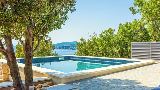 A rectangular swimming pool with a clear blue water, surrounded by a paved deck, trees, and a view of the sea and hills in the background. - Villa Turquoise Breeze (Novi Vinodolski)