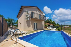 Une piscine avec des chaises longues, un balcon équipé de climatiseurs et un ciel d’un bleu éclatant.