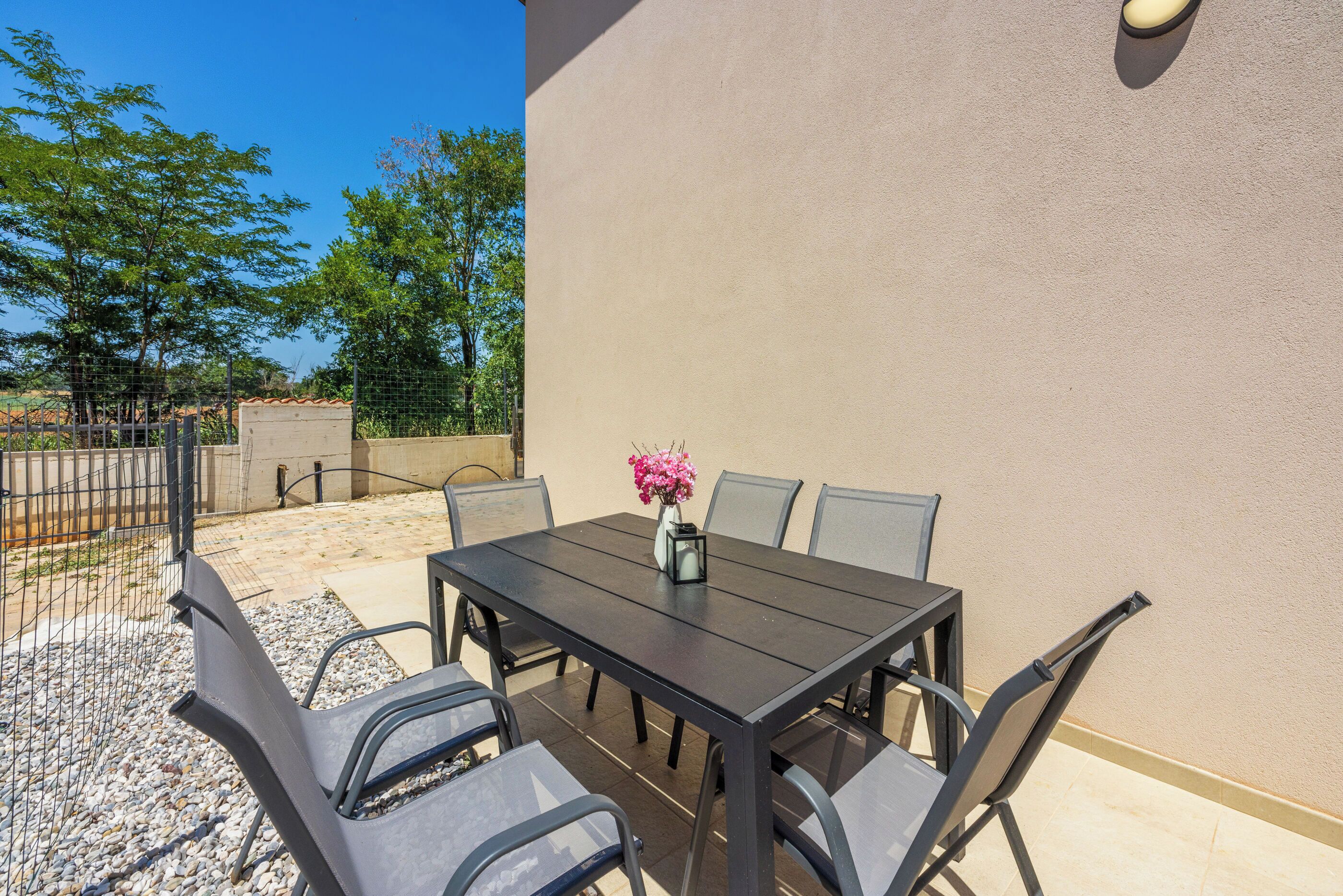 Un patio al aire libre con una mesa de madera oscura y seis sillas, un jarrón con flores y una vista de árboles y una cerca al fondo.