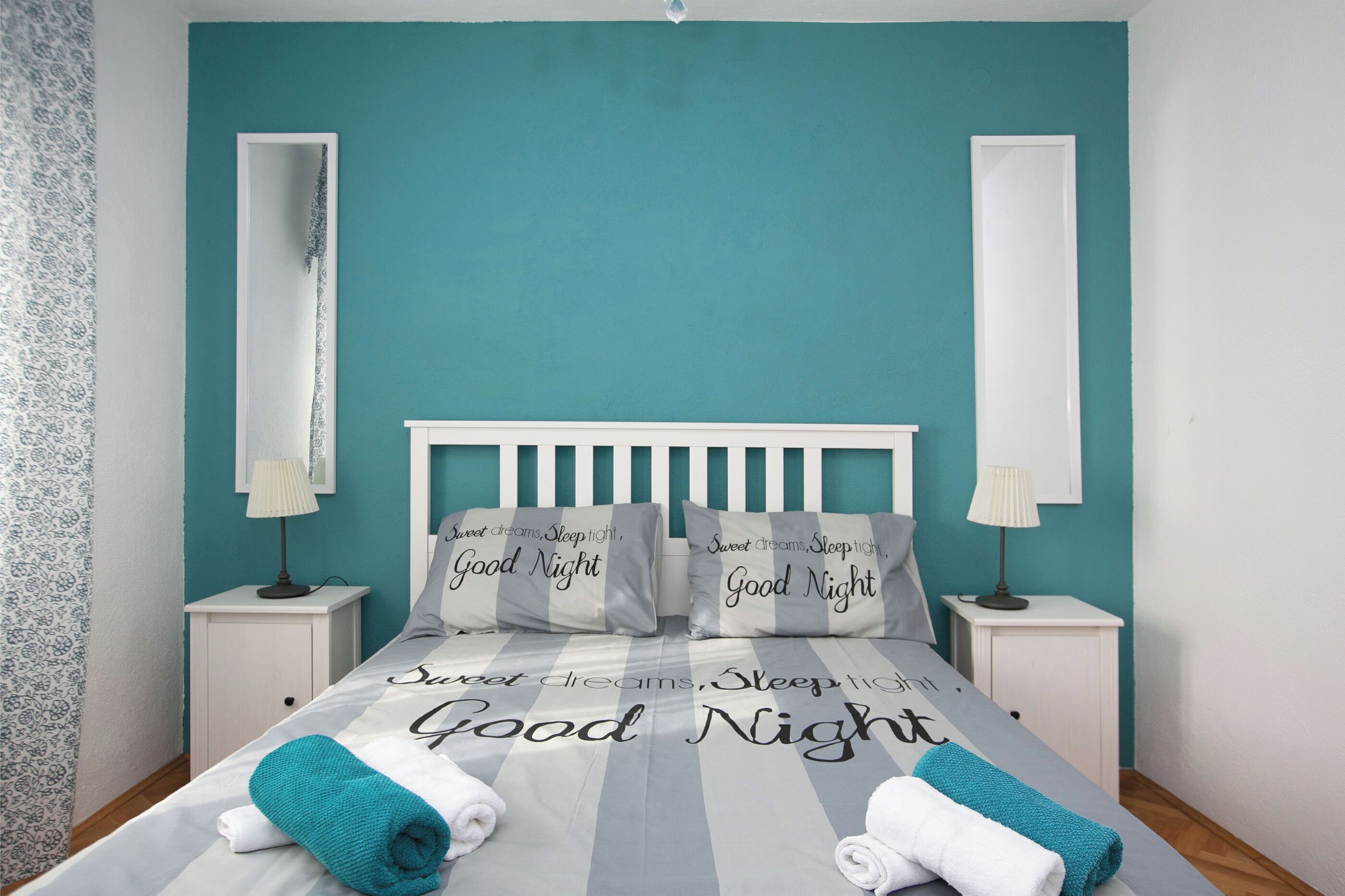 Una cama bien tendida con almohadas y una colcha, dos mesitas de noche con lámparas, y un espejo grande en la pared.