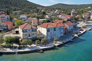 A coastal town with buildings along a waterfront, featuring balconies, a clear blue sea, and a hilly backdrop. - Apartments Tiho II (Marina)