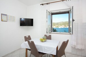 Un comedor con una mesa, sillas, un frutero, un televisor y una ventana que ofrece vistas a un pueblo costero.