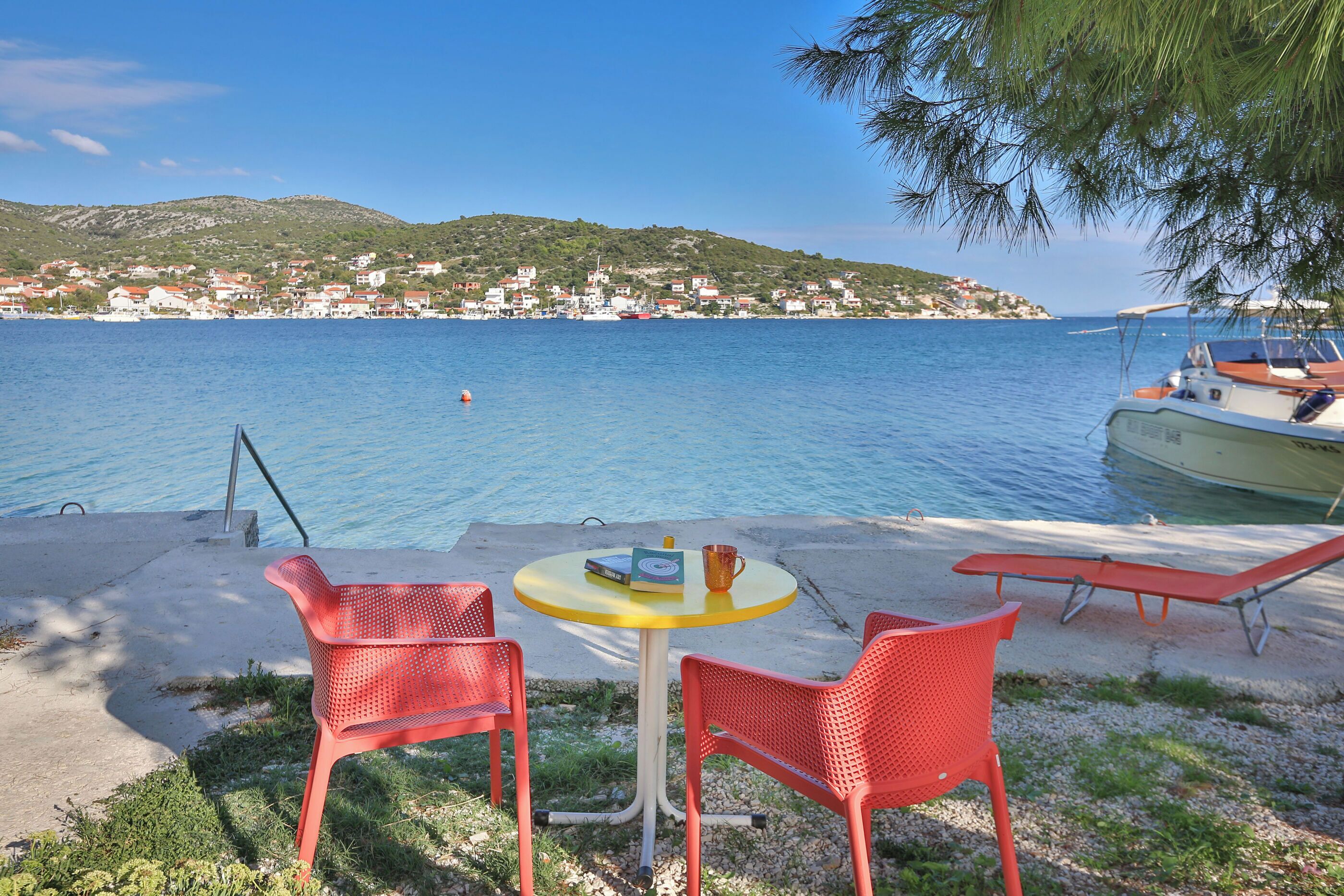 Eine Seeterrasse mit rundem Tisch und zwei roten Stühlen, mit Blick auf ein ruhiges Meer und ein Küstendorf.