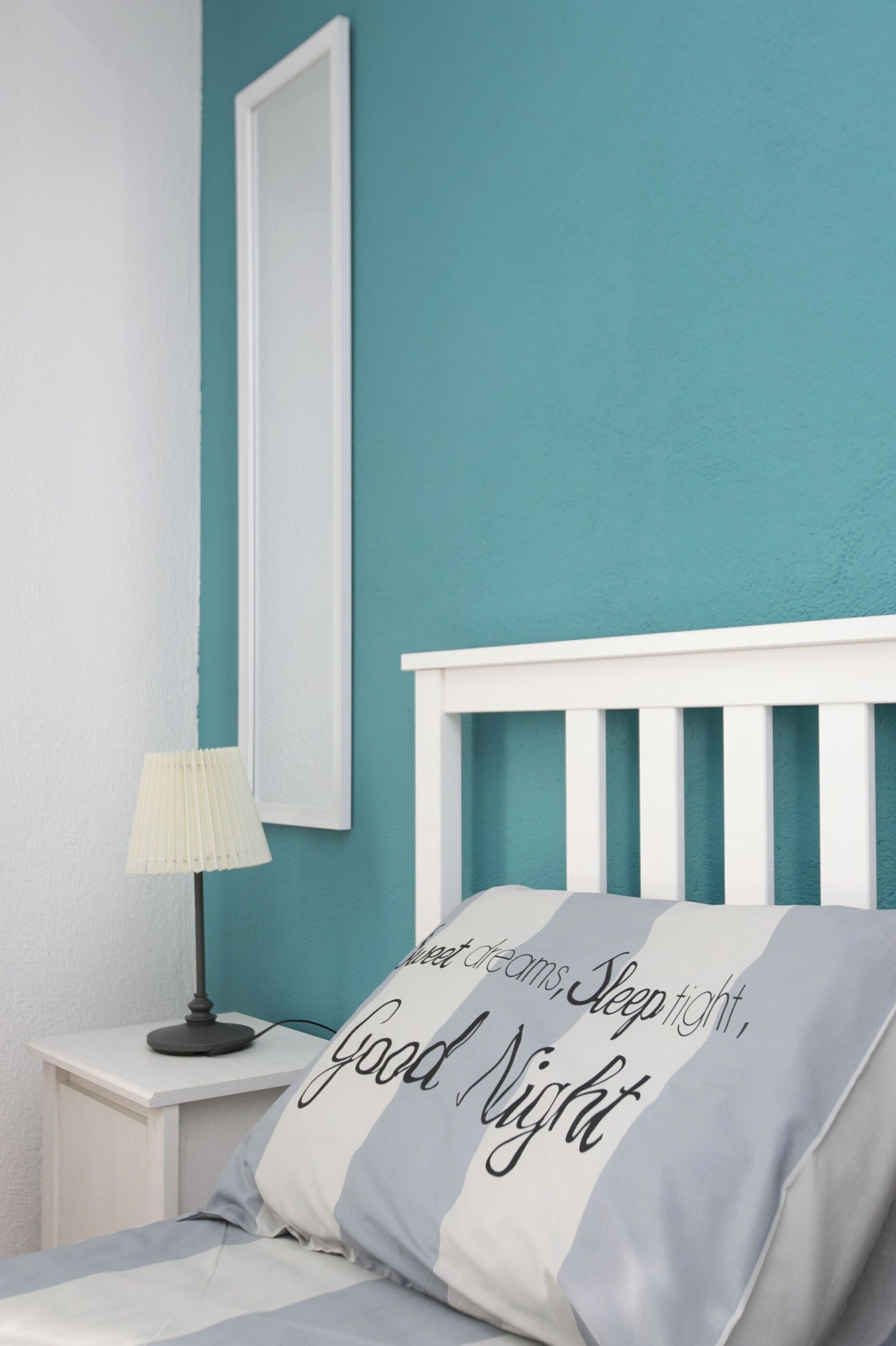 A bedroom with a teal wall, white headboard, and a bedside table with a lamp. A pillow with the text "Good Night" is visible.