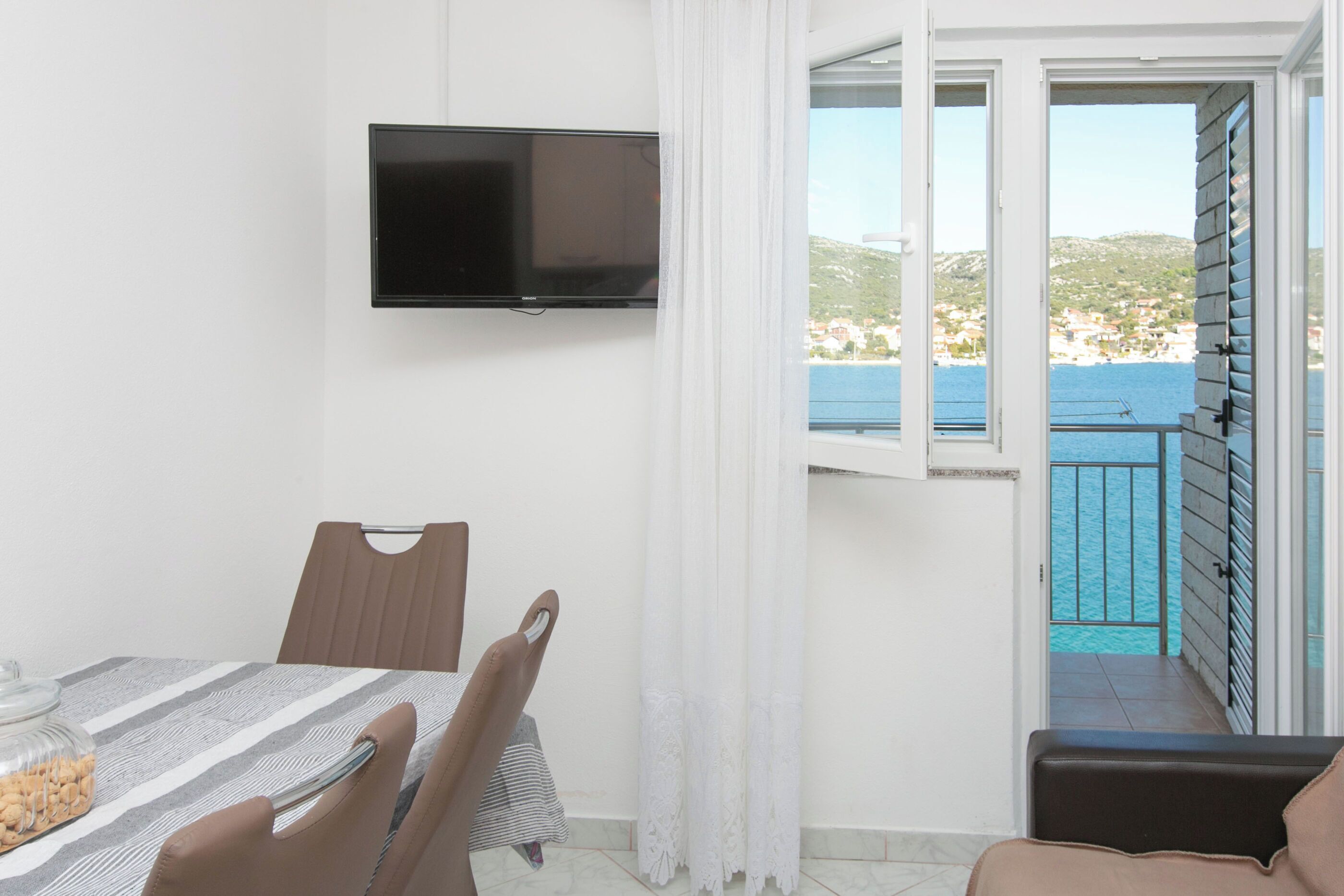 Une pièce avec un téléviseur fixé au mur, une table à manger avec des chaises, et une vue sur la mer par une porte ouverte.