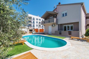 Una piscina circondata da un solarium in pietra, una sedia a sdraio in legno e un piccolo giardino con alberi di ulivo.