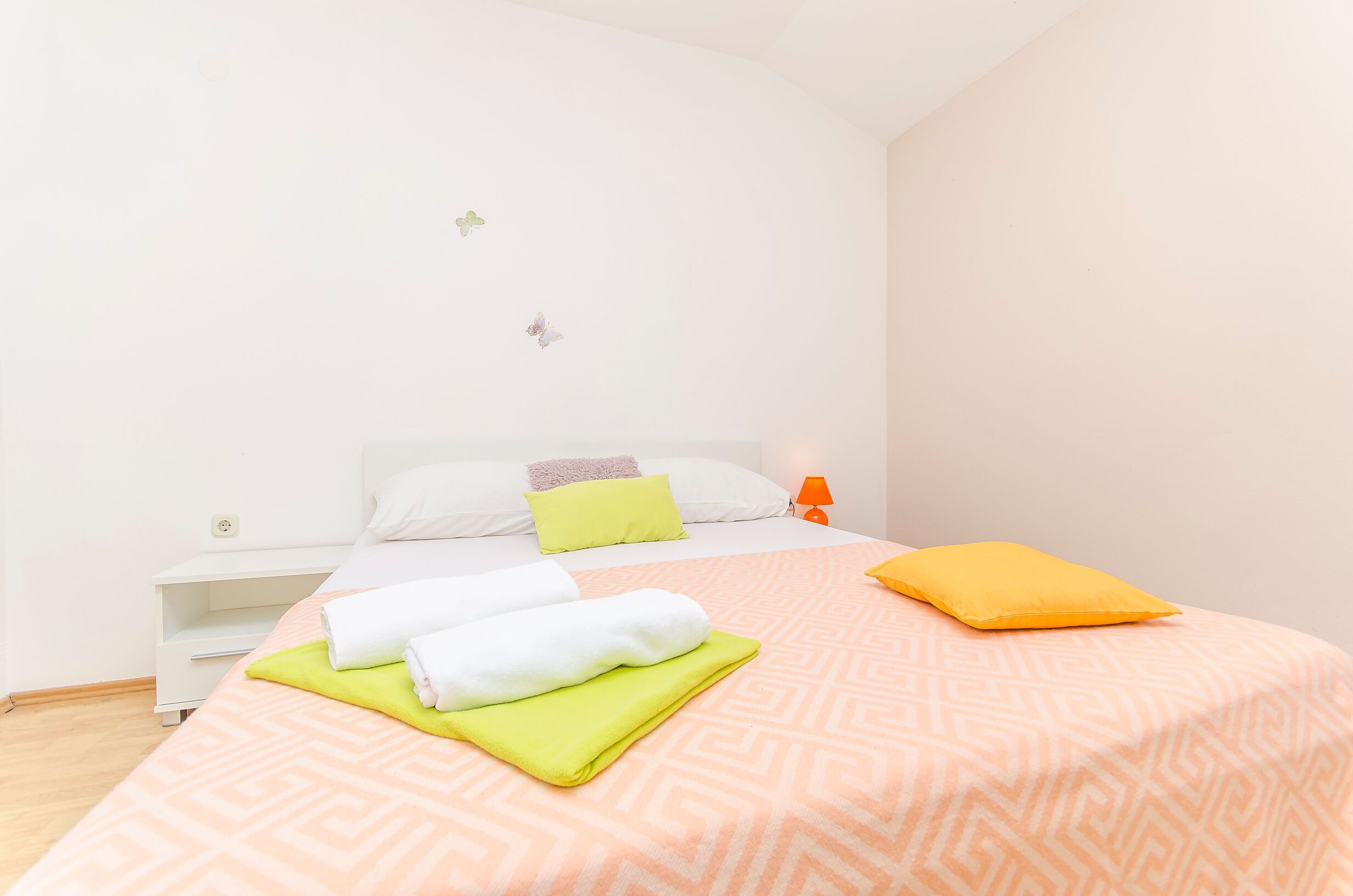 Una cama bien hecha con un edredón estampado, una mesita de noche con una lámpara y una pared decorada con adhesivos de mariposas.