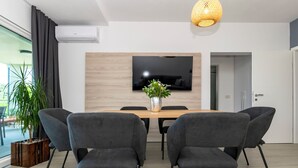Un comedor moderno con una mesa de madera, seis sillas grises tapizadas, un televisor de pantalla plana grande montado en la pared y una planta en maceta a la izquierda.