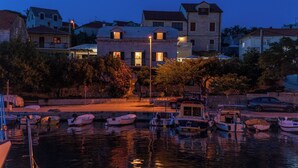 Um imóvel à beira-mar com várias embarcações atracadas, uma rua em primeiro plano e edifícios com janelas iluminadas.