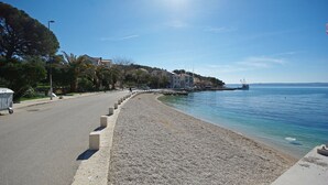 Uma estrada costeira com uma praia de seixos, um mar azul límpido e edifícios ao longo da costa.