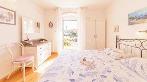 A bedroom with a bed, a chair, a dresser, a mirror, and a view of the outside.