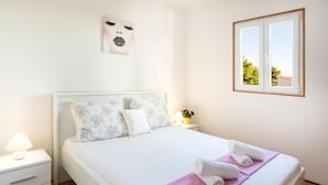 Ein ordentlich bezogenes Bett mit floralen Kissen, einer Nachttischlampe und einem Fenster mit Blick auf Grünflächen.