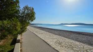 Ein gepflasterter Weg entlang eines Kiesstrandes, der zu einer ruhigen Wasserfläche führt, mit Bergen im Hintergrund und einem klaren Himmel.