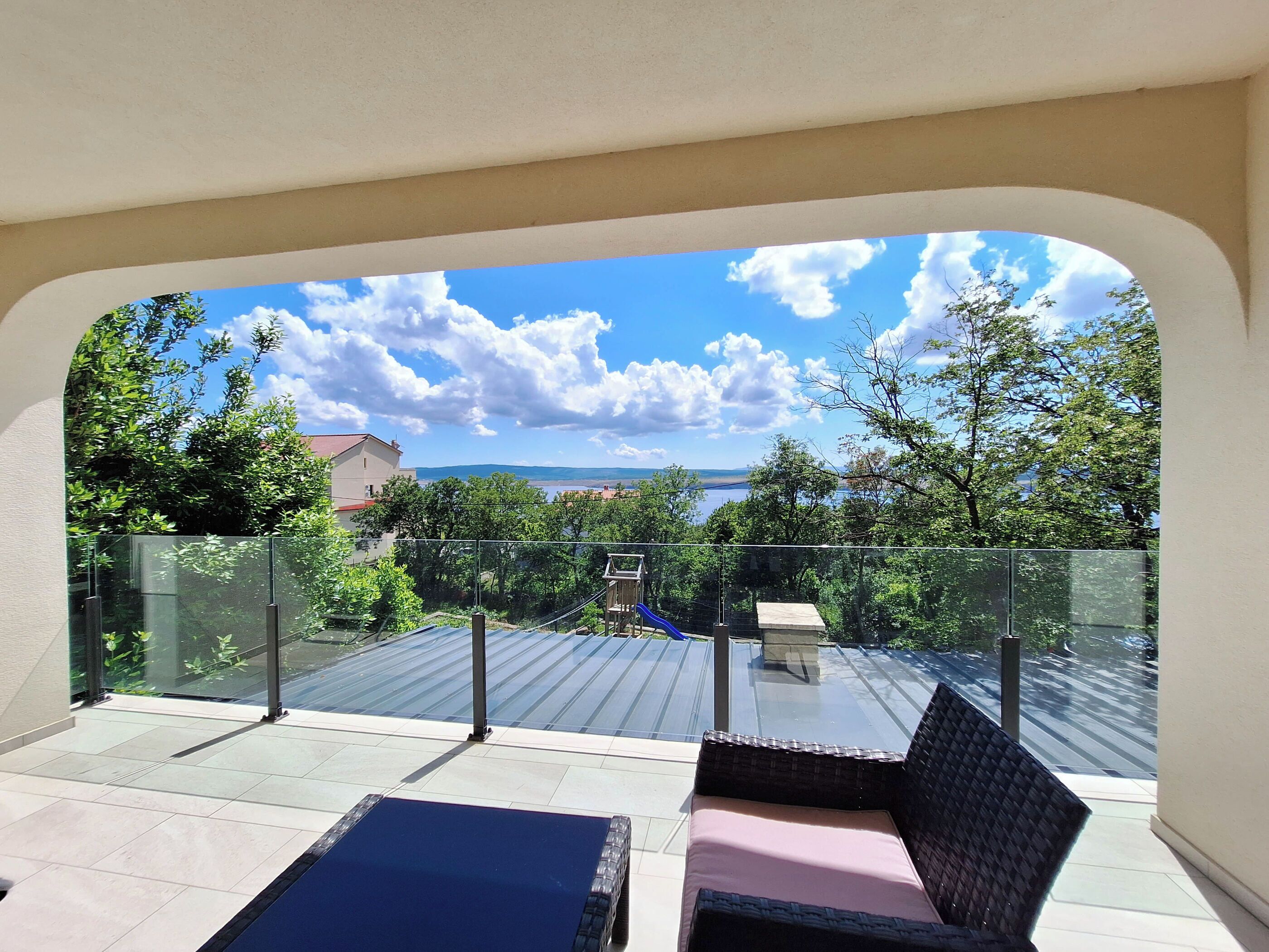 Un balcon doté d’une balustrade en verre, un canapé bleu et une vue sur un lac et des bâtiments au loin.
