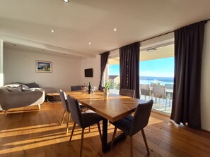 A modern living room with a wooden dining table, grey sofa, and a large window with a view of the sea. - Apartments Zoran (Crikvenica)