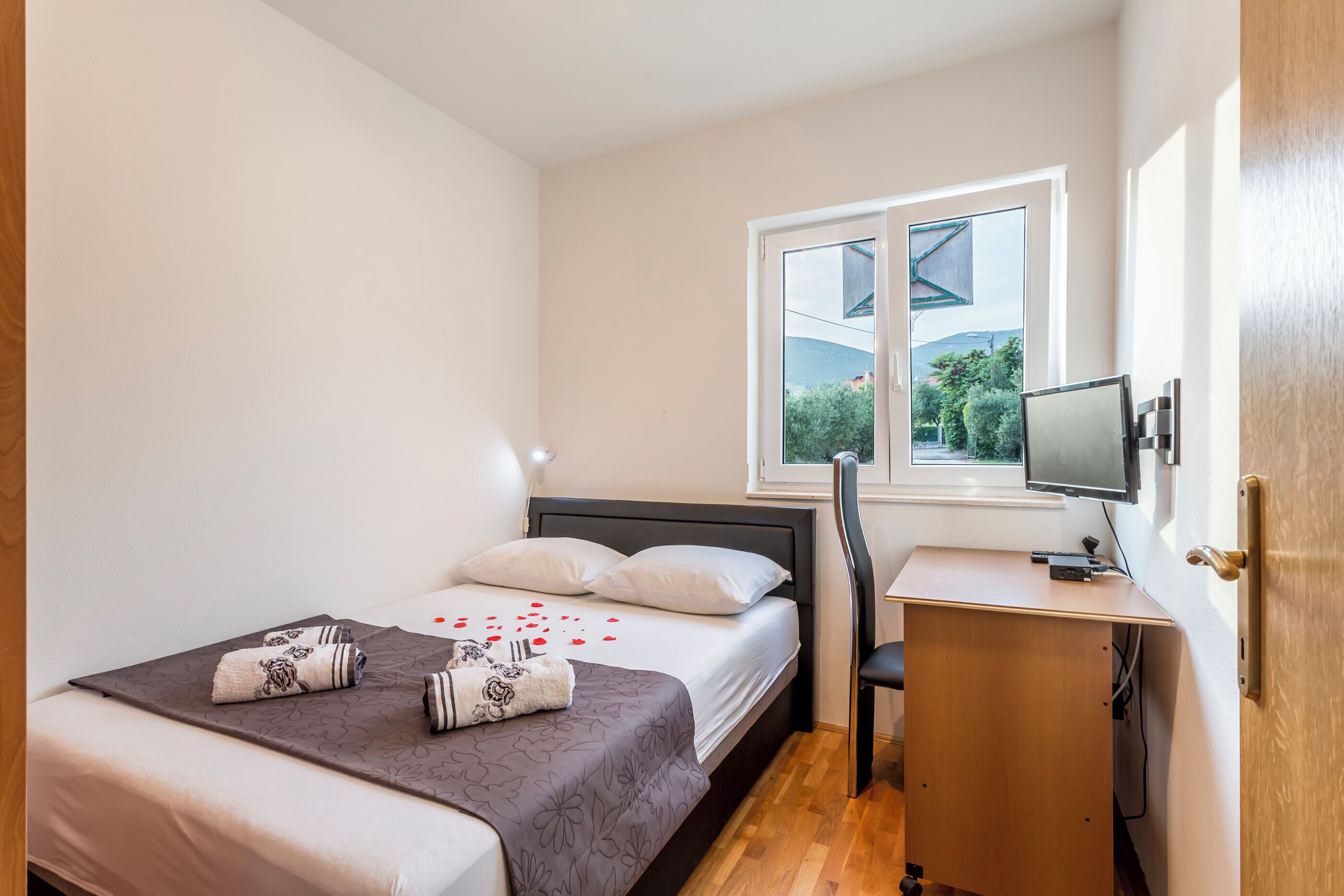 A bedroom with a bed, a desk, a chair, a TV, and a window with a view of greenery.