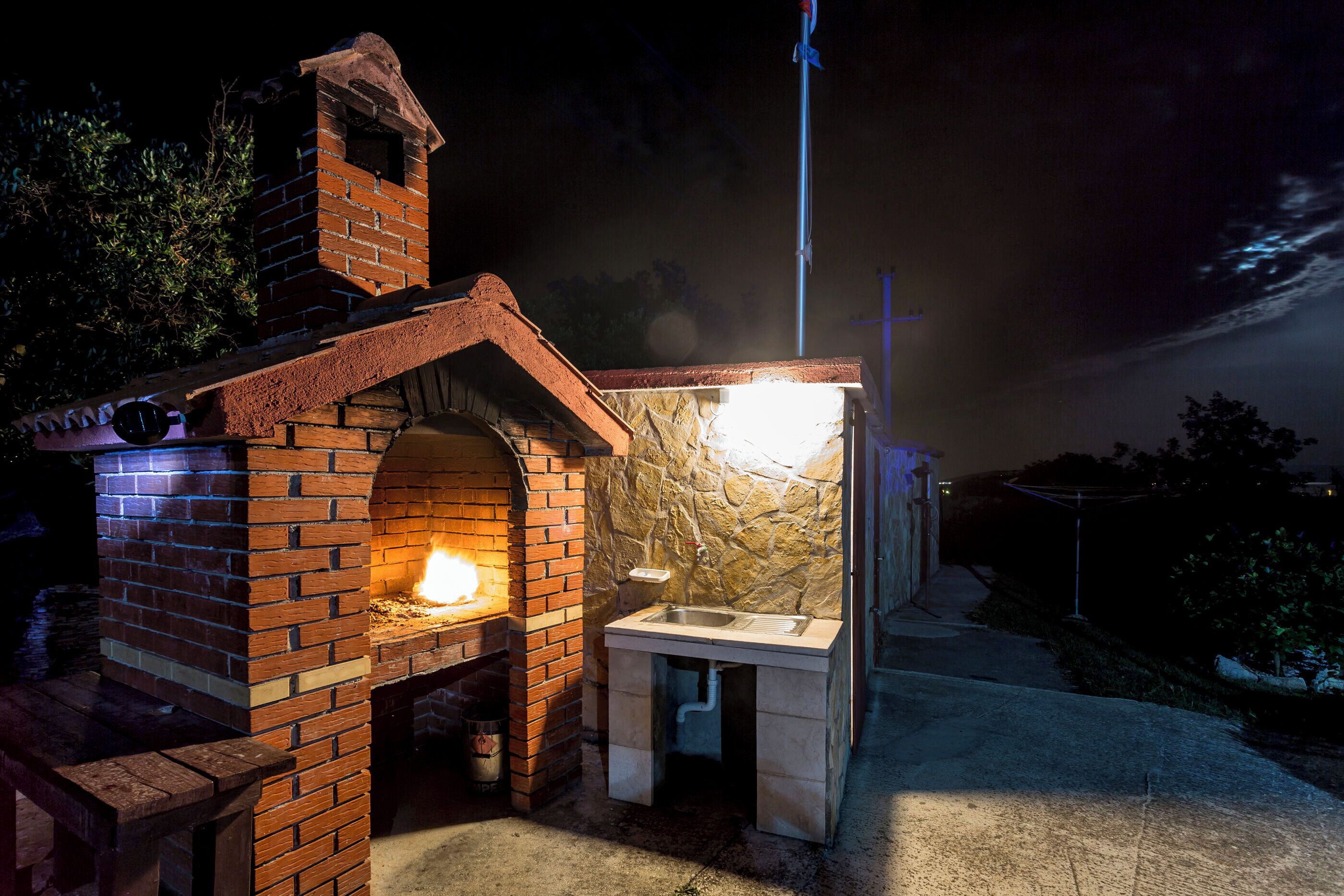 A brick-built outdoor kitchen with a stone wall and a sink, surrounded by a concrete area.