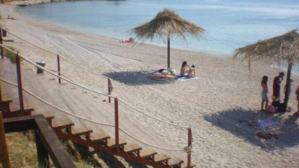 Ein Strand mit einer Holzzaun, reetgedeckten Sonnenschirmen und freiem Blick auf das Meer und die dahinterliegenden Berge.