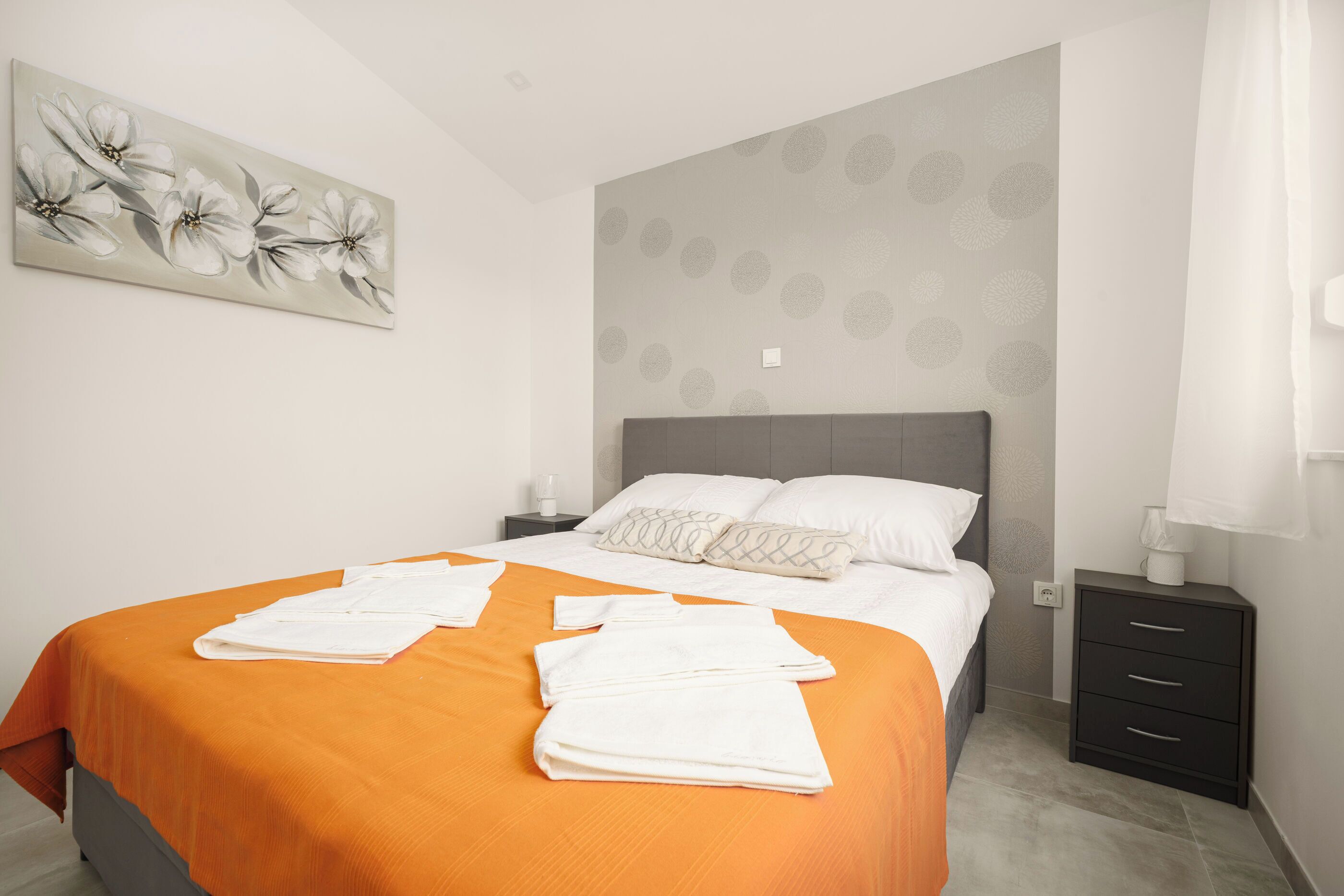 Ein Schlafzimmer mit orangefarbener Bettdecke, grauem Kopfteil, einem Nachttisch mit Lampe und einer Wand mit einem floralen Gemälde.