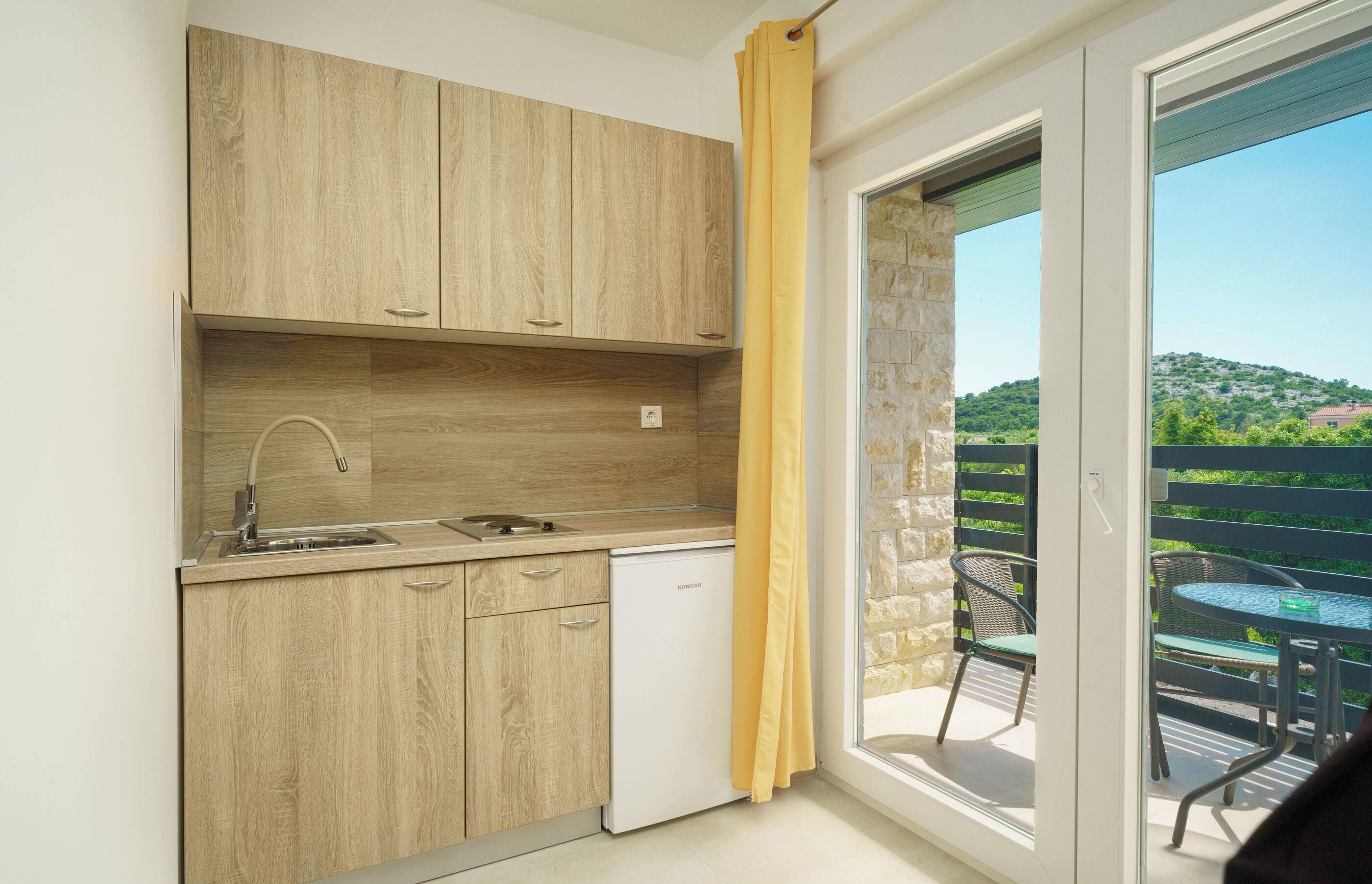 A compact kitchen with wooden cabinets, a built-in dishwasher, and a window overlooking a balcony with outdoor furniture.