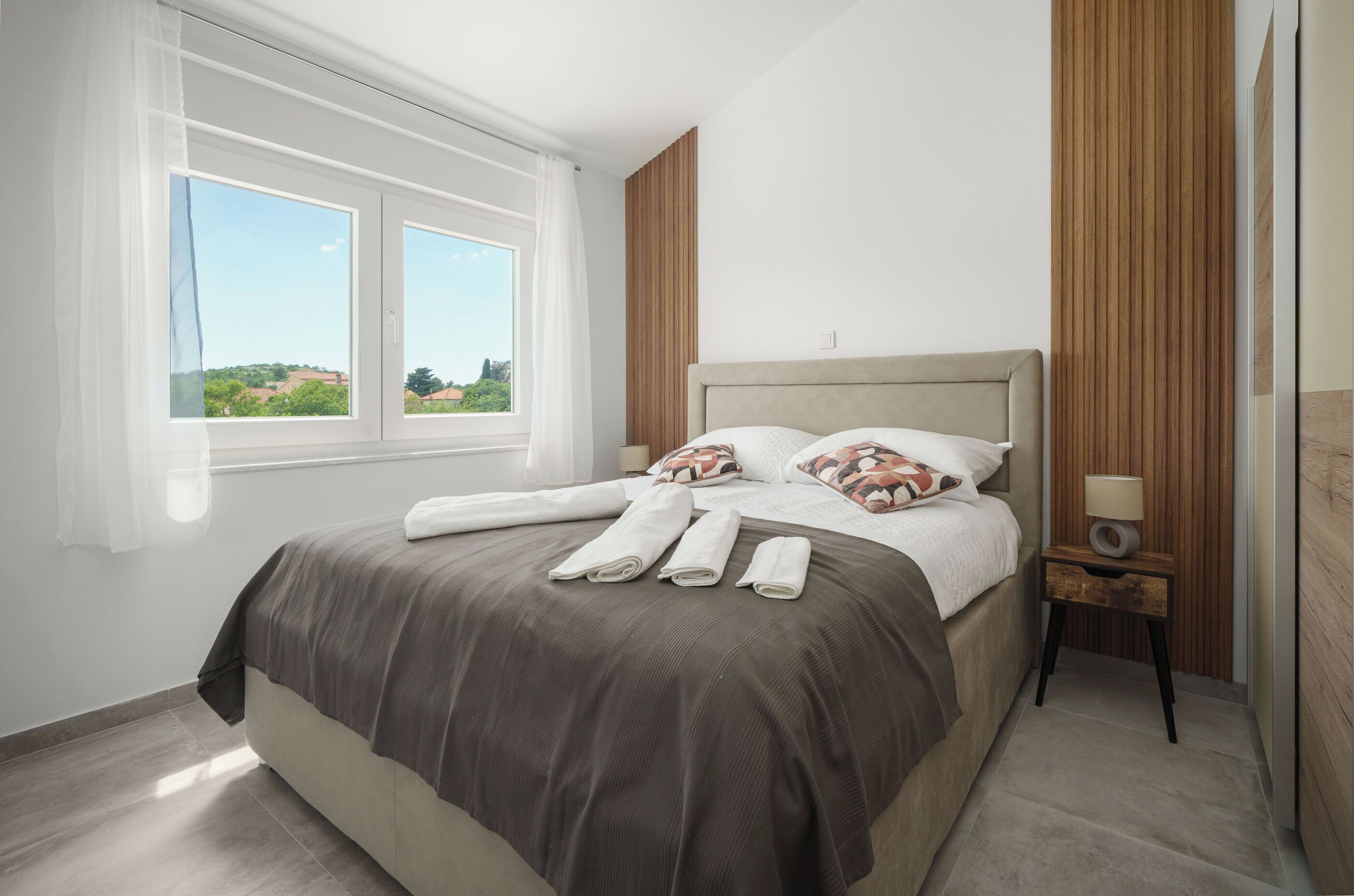 A neatly made bed with a brown bedspread and pillows, a bedside table with a lamp, and a window with a view of greenery.