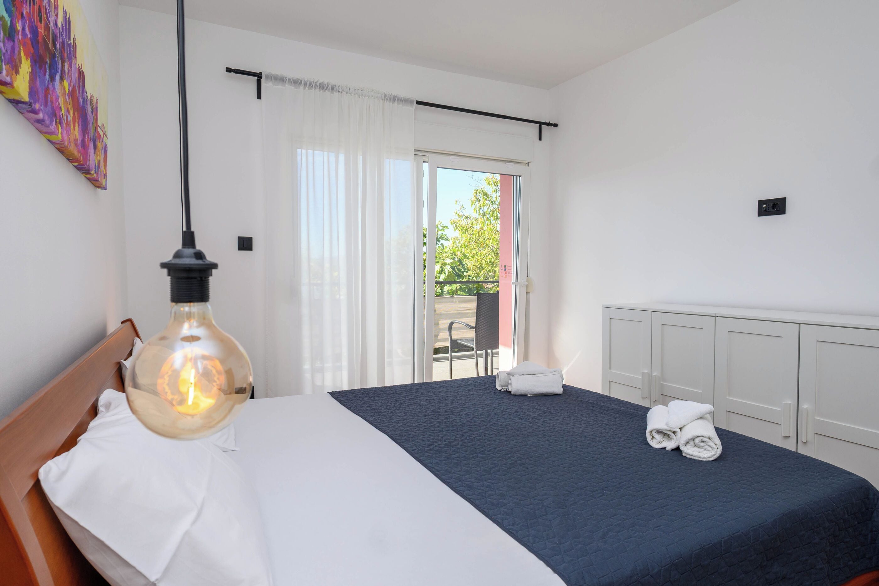 A hotel room with a bed, a hanging light fixture, a view through the window, and a radiator.